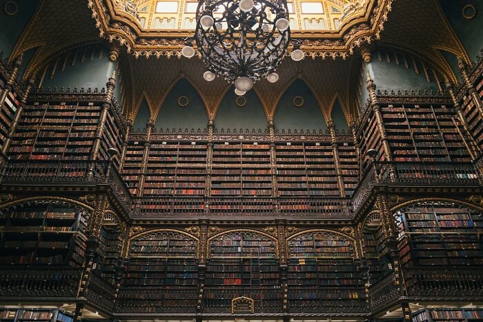 Prächtiger, grandioser Bibliotheksinnenraum mit hoch aufragenden Bücherregalen und filigranen Golddetails unter einem dekorativen Kronleuchter.